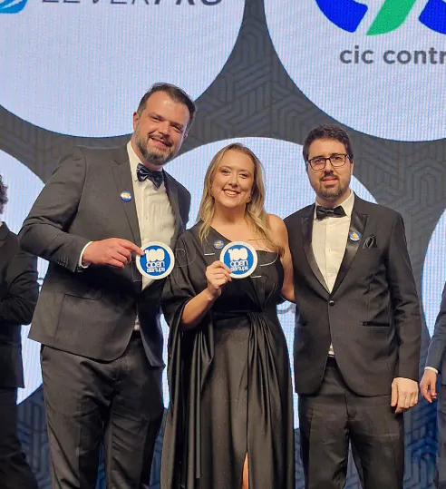 Três pessoas vestidas de gala posam sorridentes em um evento de premiação, segurando placas com a marca "100 Open Startups".